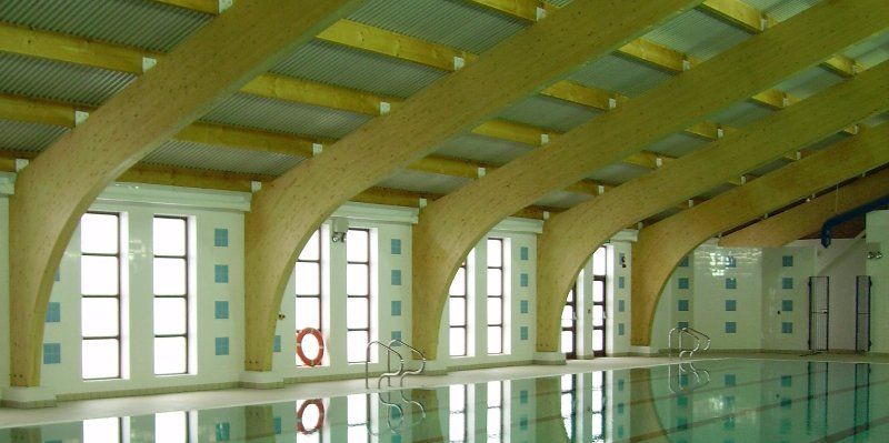 A large indoor swimming pool with corrugated roof and timber structures extending across the ceiling from one side of the pool to the other